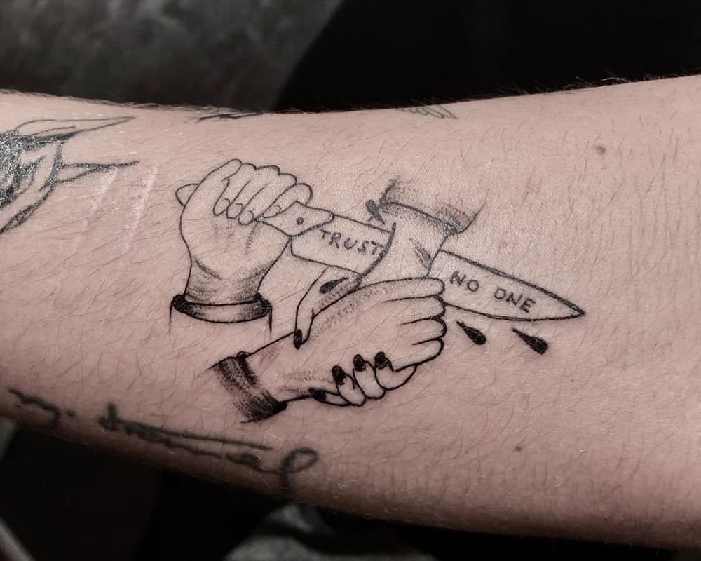 A tattoo of one hand stabbing the other with a knife that says trust no one