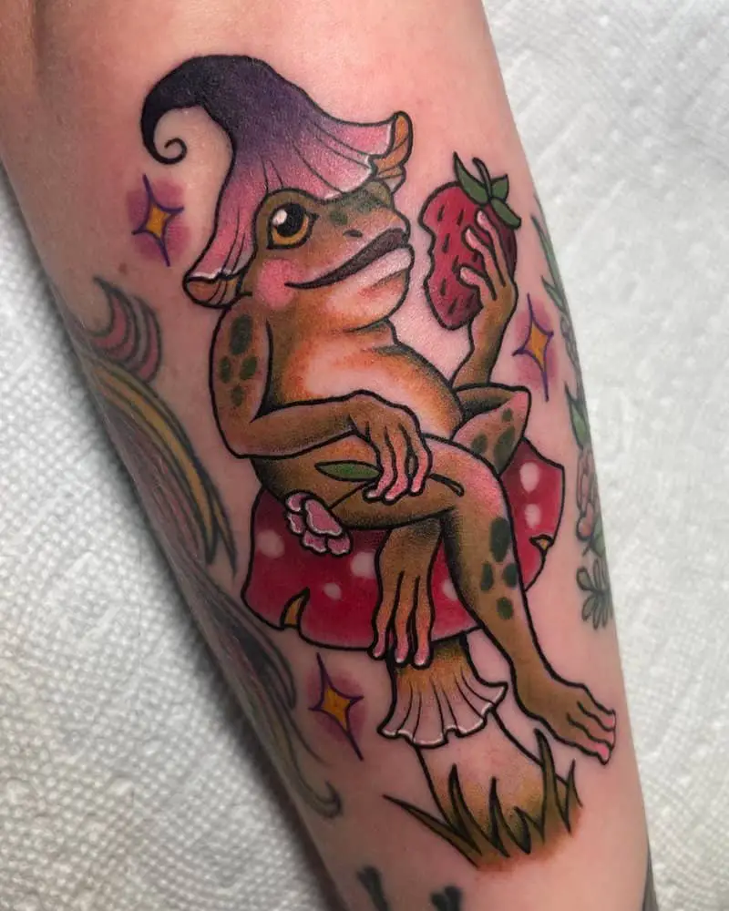 Tattoo of a frog wearing a flower hat sitting on a mushroom and eating strawberries