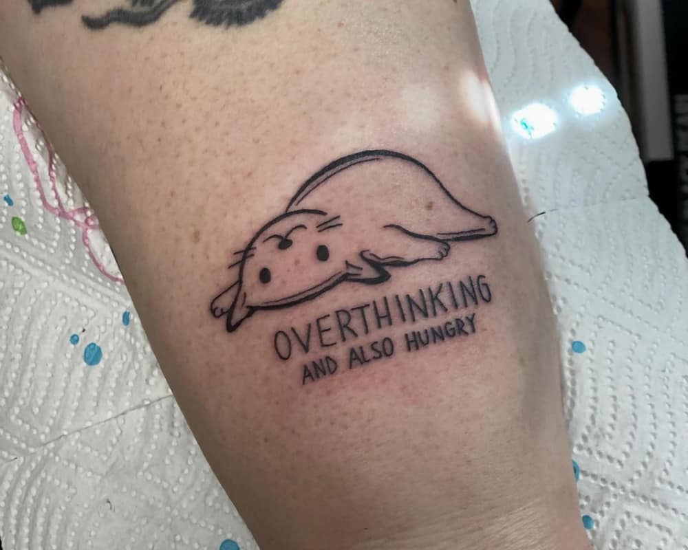 a tattoo of a lying cat and the inscription overthinking and also hungry